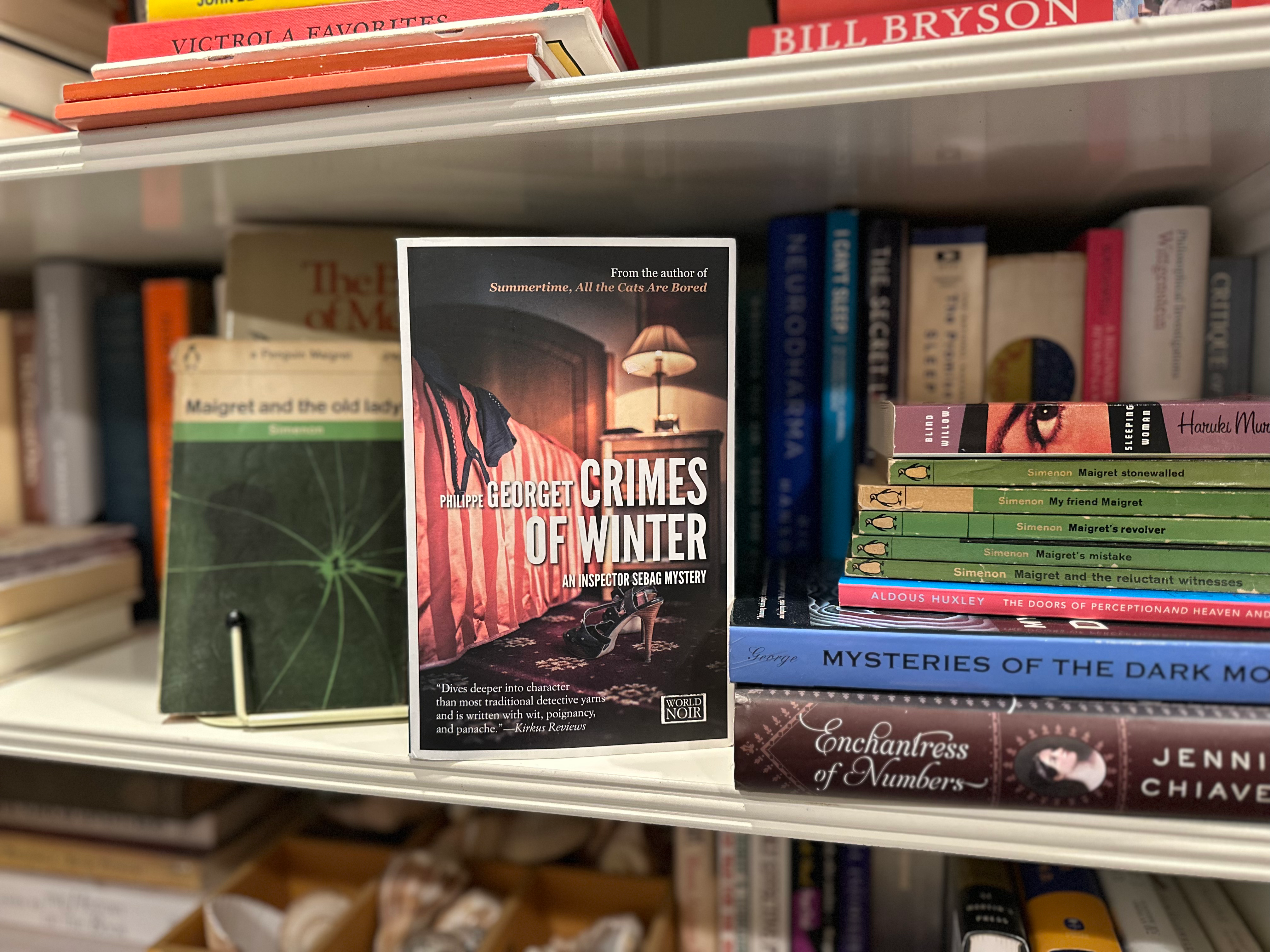 Photo of 'Crimes of Winter' novel propped up on a white bookcase, with green vintage Penguin editions of Maigret novels surrounding it